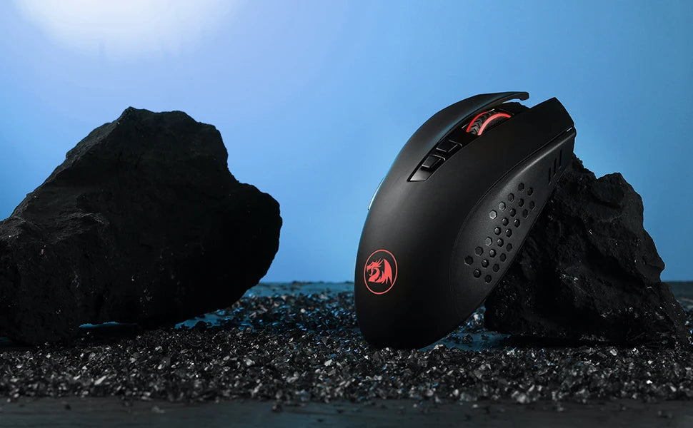 A black Redragon wireless gaming mouse is placed on dark gravel next to large rocks under a blue sky.
