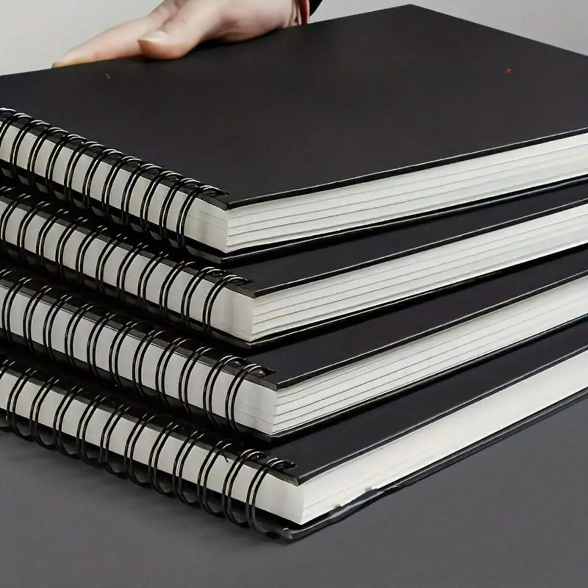 A stack of black and white spiral-bound sketchbooks with white pages is held by a hand.