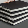 A stack of black and white spiral-bound sketchbooks with white pages is held by a hand.
