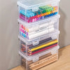 A stack of clear plastic storage boxes filled with various colored markers and pencils for office and craft supplies.