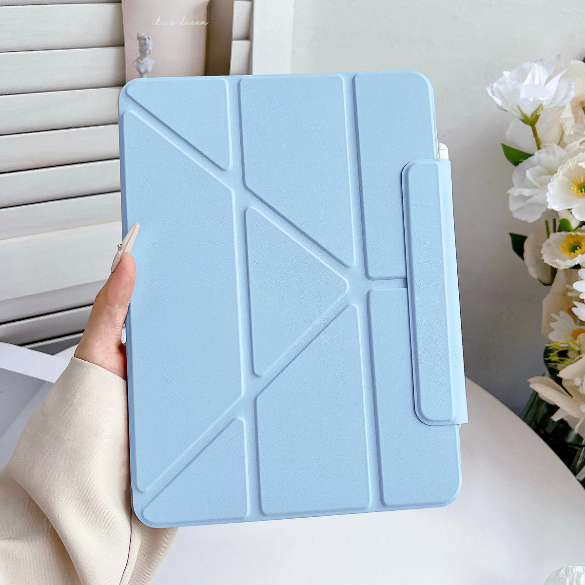 Una funda protectora de imitación de cuero azul claro para un iPad sostenida por una mano, apoyada sobre una superficie blanc