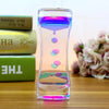 A glass hourglass timer filled with colorful liquid, resting on a wooden surface next to stacked books and pink flowers.