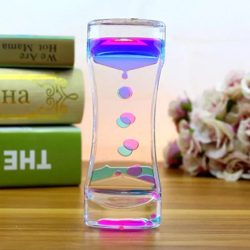 A glass hourglass timer filled with colorful liquid, resting on a wooden surface next to stacked books and pink flowers.