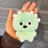 A hand holds a light green plush toy shaped like an animal with white ears and a beaded necklace.