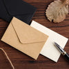 Una pila de sobres de invitación de papel kraft y una carta colocada sobre una superficie de madera oscura junto a una hoja s