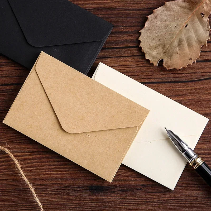 Una pila de sobres de invitación de papel kraft y una carta colocada sobre una superficie de madera oscura junto a una hoja s