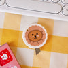 A round, light brown cat face stamp or mini cutter is placed on a yellow and white checkered surface next to a pink item.
