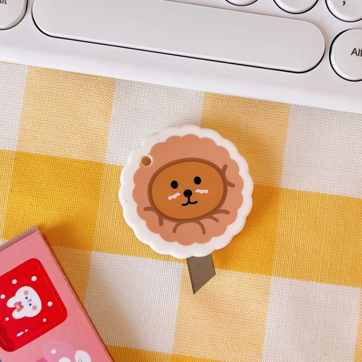 A round, light brown cat face stamp or mini cutter is placed on a yellow and white checkered surface next to a pink item.