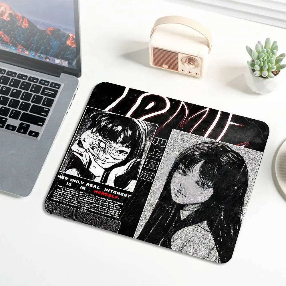 A black mouse pad featuring a Junji Ito Tomie design with stitched edges, placed next to a laptop and a small tin.
