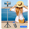A woman holds a selfie stick tripod with a camera and Bluetooth remote, set up outdoors near the sea.