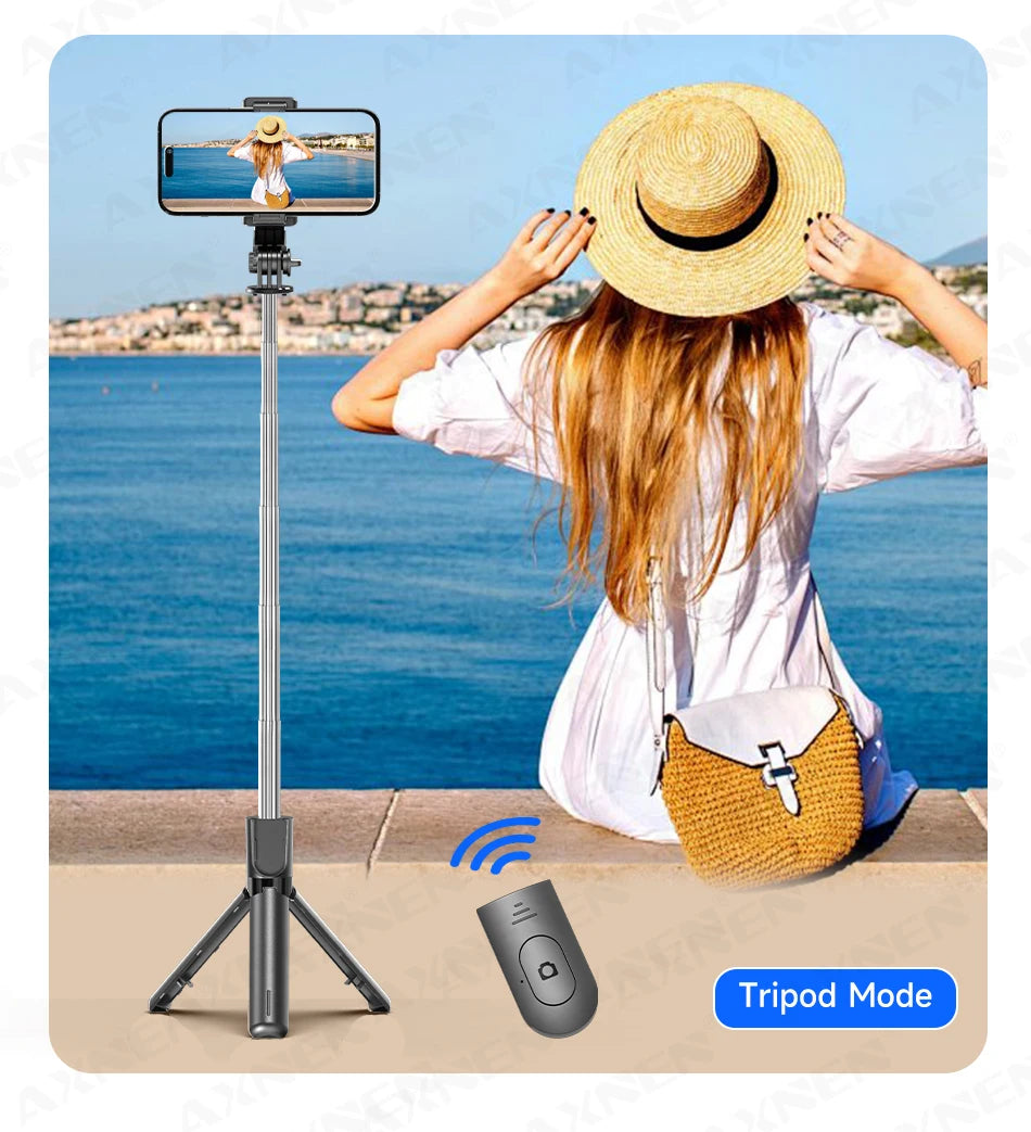 A woman holds a selfie stick tripod with a camera and Bluetooth remote, set up outdoors near the sea.