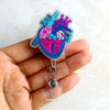 A hand holds a shiny enamel badge reel featuring an anatomical diagram of the heart in blue and pink.