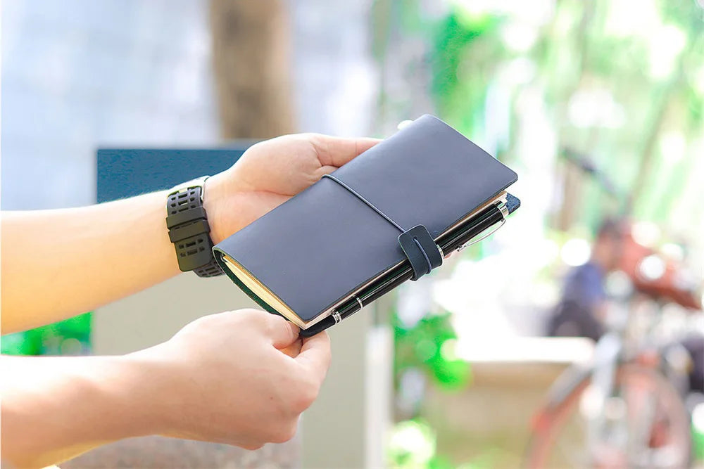 يد تمسك بمفكرة سفر جلدية رمادية داكنة قابلة لإعادة التعبئة أمام خلفية خضراء خارجية ضبابية.