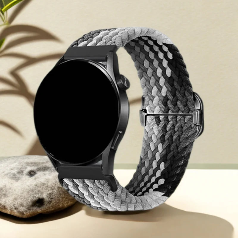A black smartwatch is shown on a gray patterned nylon watch strap, resting on a light surface.