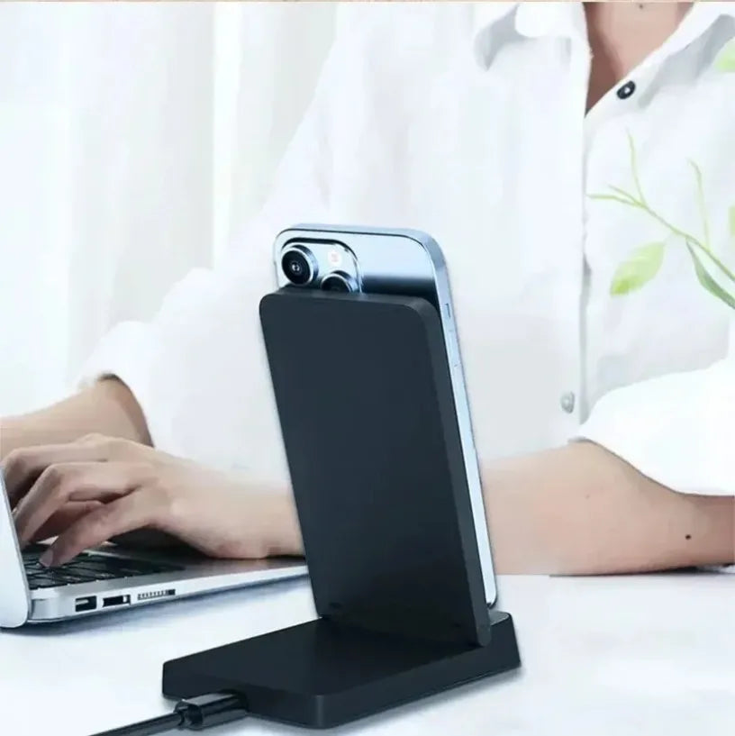 A black magnetic foldable wireless charging stand is shown next to a laptop on a white surface.