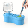 A hand is using a blue paint brush to clean a clear glass jar of paint or liquid inside a blue plastic container.