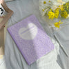 A purple composition book cover with a heart design lying on a white cloth next to a pencil holder and yellow flowers.