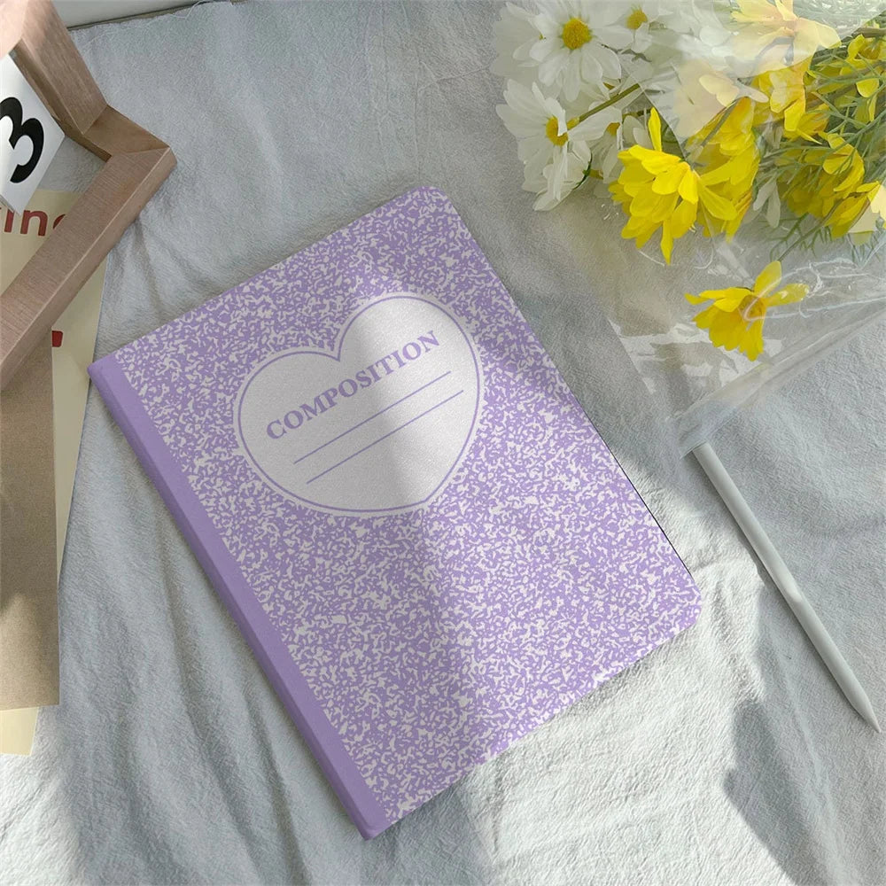 A purple composition book cover with a heart design lying on a white cloth next to a pencil holder and yellow flowers.