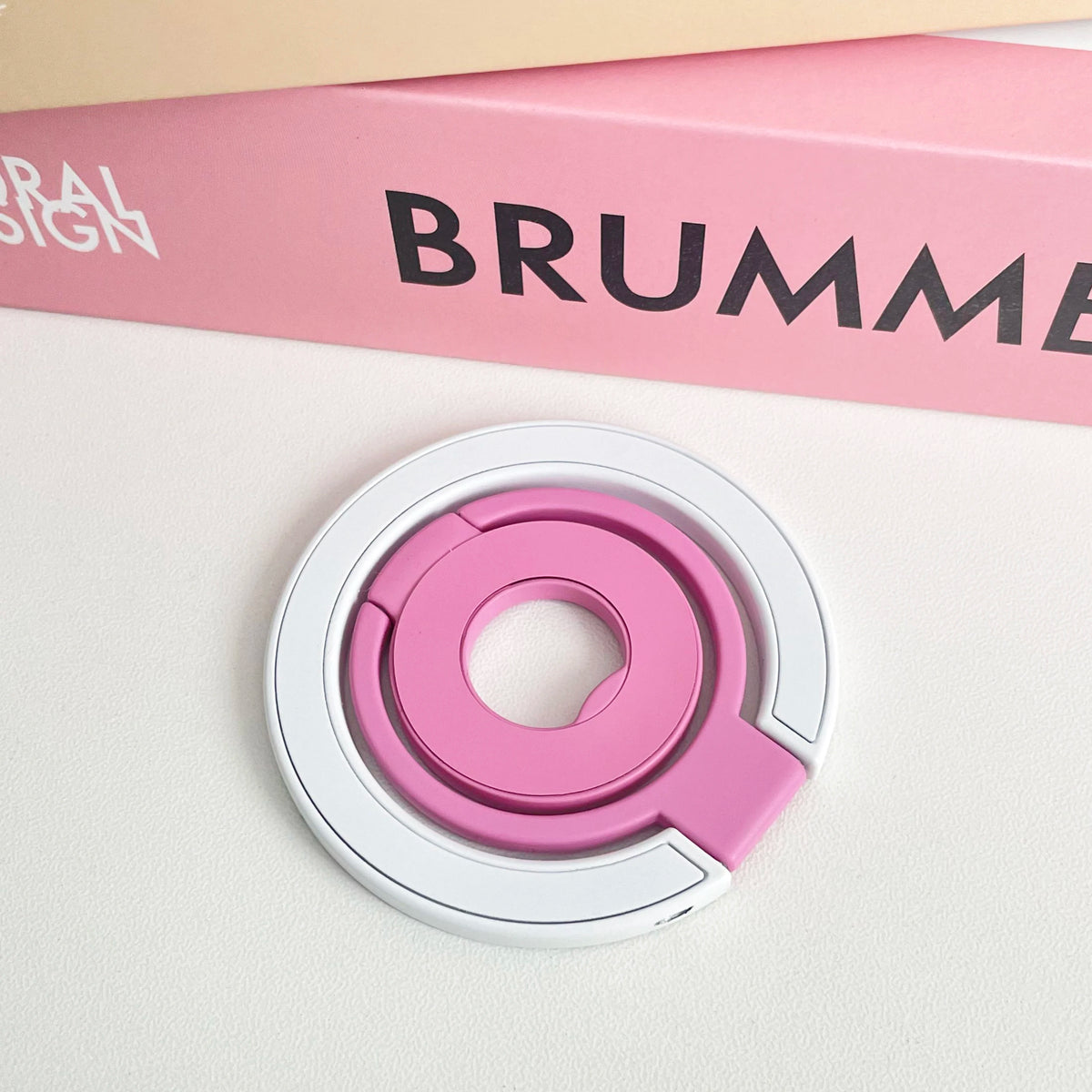 A pink magnetic phone grip and stand with a foldable ring is shown on a white surface.