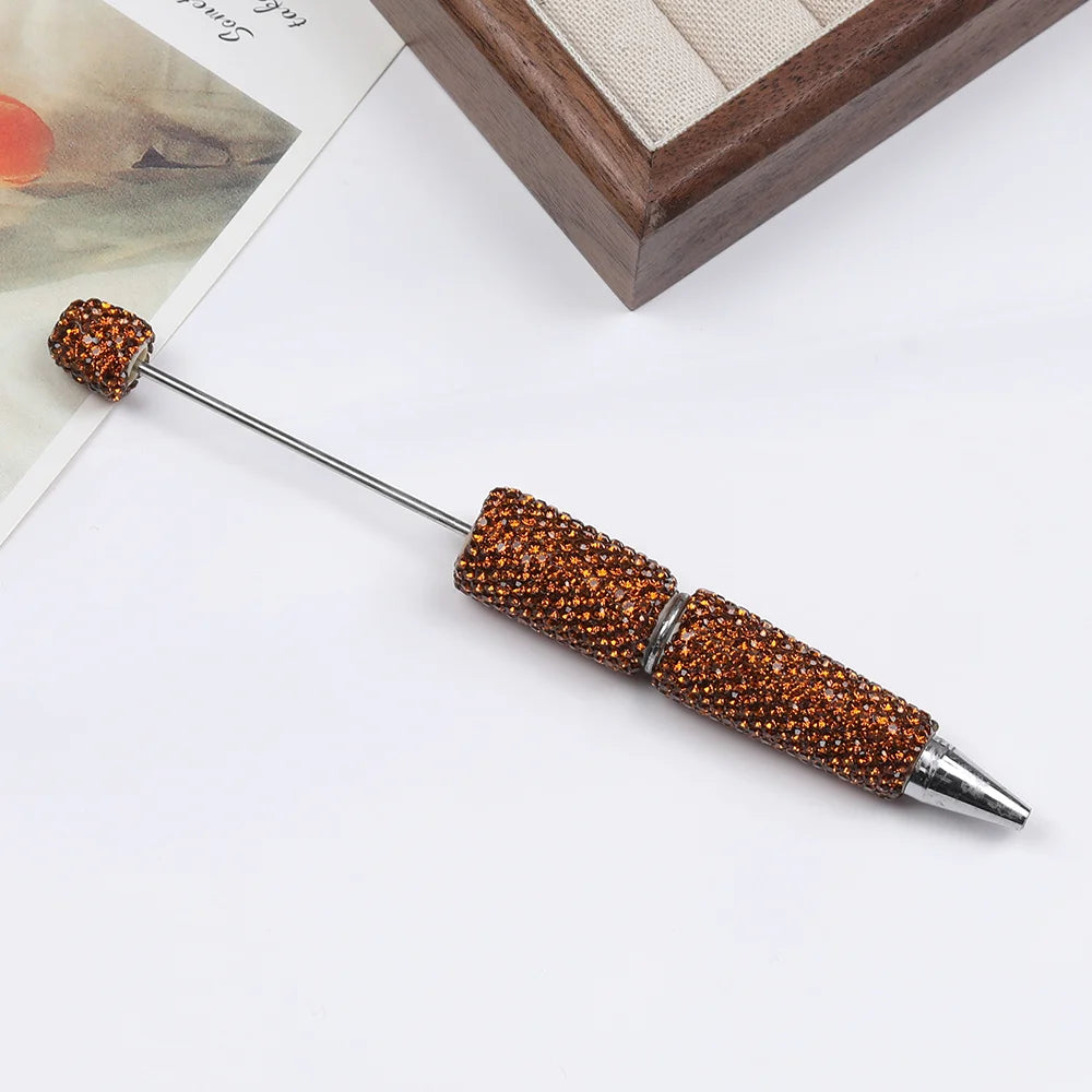 A long, silver pen accessory with a gold-colored beaded tip rests on a white surface next to a wooden frame.