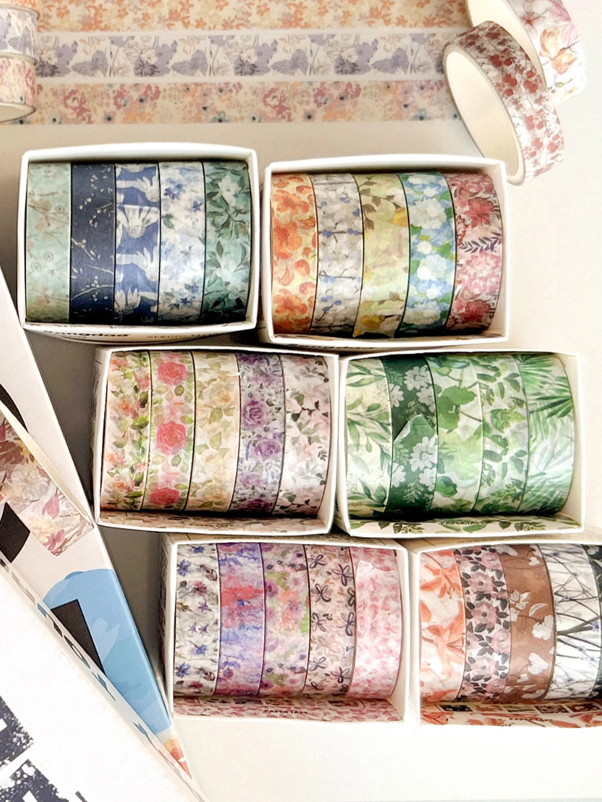 A collection of five rolls of floral patterned washi tape in various colors and designs displayed in white containers.