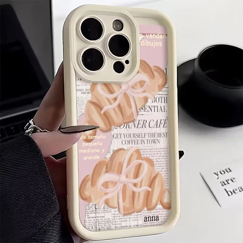 A beige phone case with a croissant newspaper pattern featuring illustrations of croissants and text.