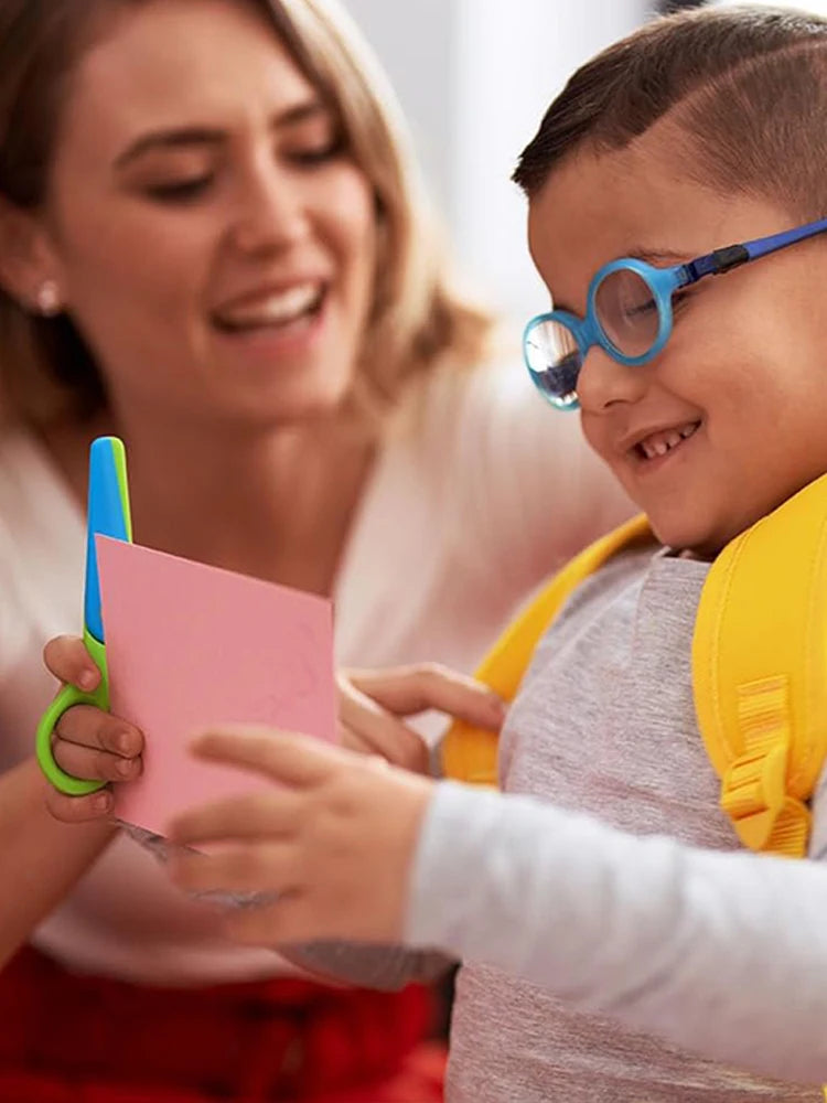 A young boy is using two-tone plastic scissors to cut paper with the help of an adult.