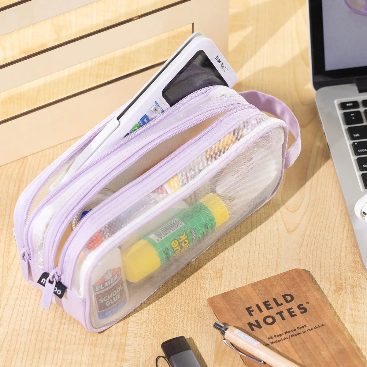 A purple transparent large-capacity pencil case is shown lying on a wooden surface next to a notebook and a pen.