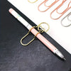 Un bolígrafo rosa con clip dorado junto a varios otros clips de bolígrafos dorados y plateados sobre una superficie negra.