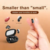 A hand holds a black wireless earbud, next to a peanut and a small charging case.