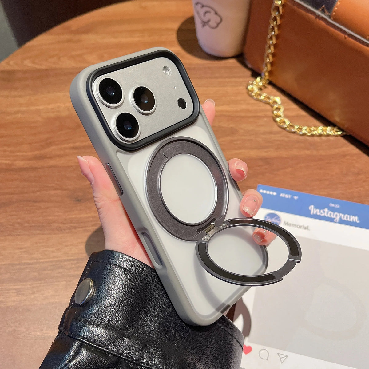 A hand holds a silver phone case with a magnetic rotating ring attached, resting on a wooden table next to an Instagram card.