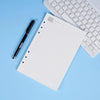 Una página de cuaderno en rejilla blanca junto a un bolígrafo negro y el teclado de una laptop plateada sobre un fondo azul c