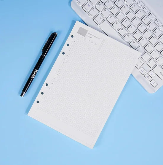 Una página de cuaderno en rejilla blanca junto a un bolígrafo negro y el teclado de una laptop plateada sobre un fondo azul c