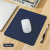 A navy blue mouse pad with a white Apple mouse placed on a wooden desk next to a laptop and a pen.