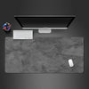 A gray overlocked mouse pad is shown with a computer monitor, keyboard, and mouse on a dark surface.