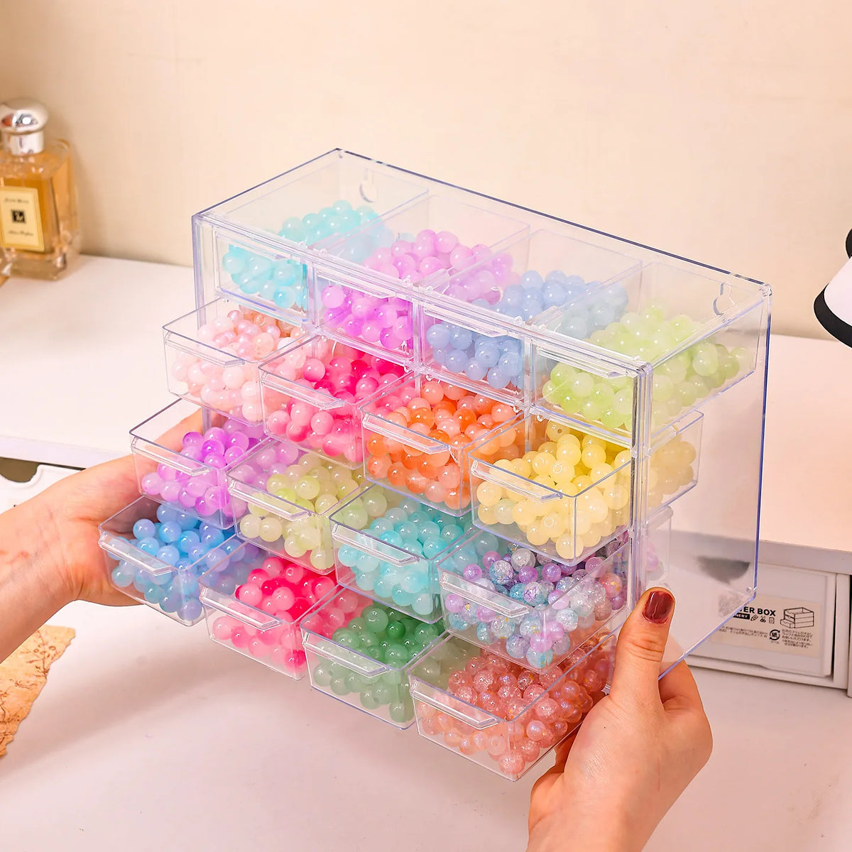 A hand holds an acrylic transparent bead storage box with multiple stackable compartments filled with colorful plastic beads.