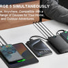 A black power bank with built-in AC plug and dual cables is shown next to several smartphones on a white desk.
