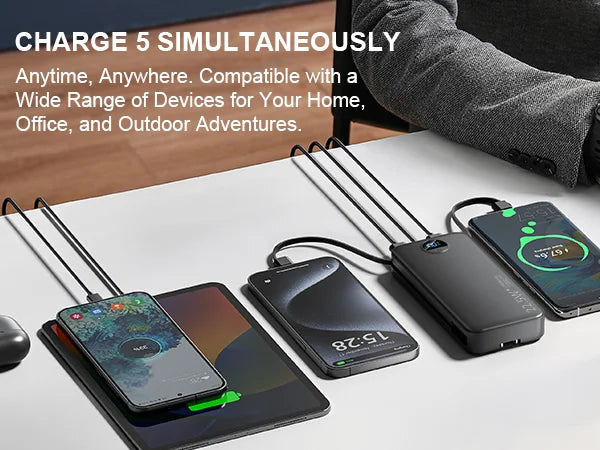 A black power bank with built-in AC plug and dual cables is shown next to several smartphones on a white desk.