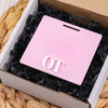 A pink acrylic badge holder with the letters OT is placed inside a cardboard box lined with black shredded paper.