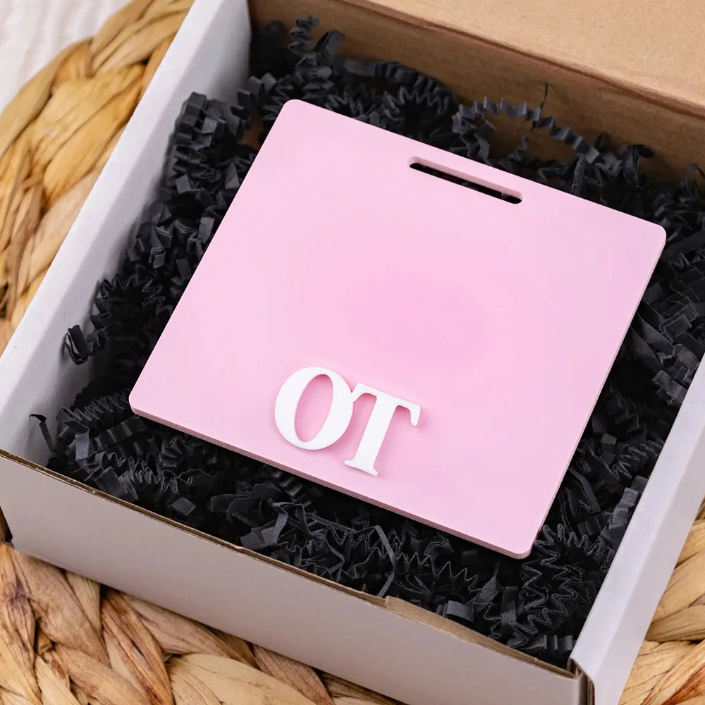 A pink acrylic badge holder with the letters OT is placed inside a cardboard box lined with black shredded paper.