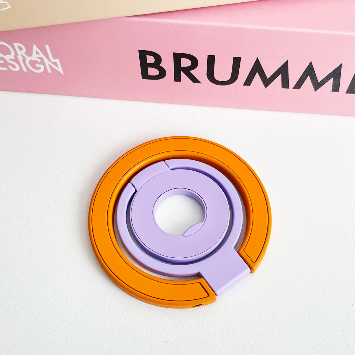 Orange and purple magnetic phone grip and stand with a foldable ring resting on a white surface.