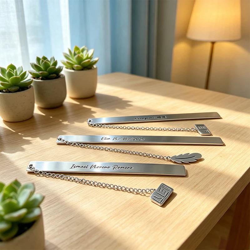Stainless steel bookmarks with engraved text displayed on a wooden table next to potted succulents.