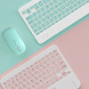 Un teclado Bluetooth blanco junto a un ratón verde azulado sobre un fondo rosa.