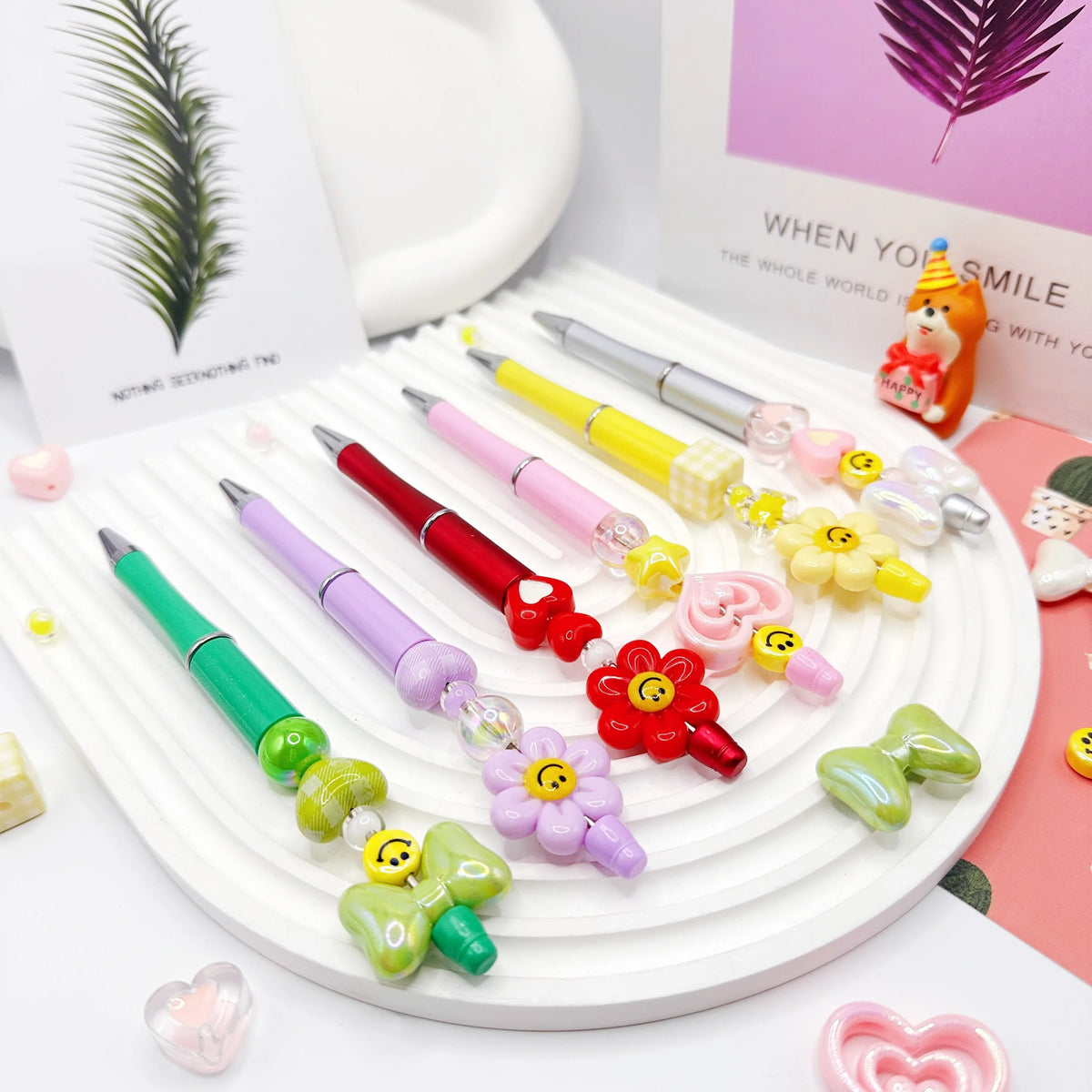 A set of colorful beaded ballpoint pens arranged on a white plate with a potted plant and a pink background.