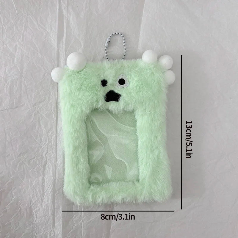 A light green plush holder shaped like a monster with white pom-poms and a clear window for a photocard.