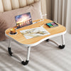 A wooden laptop desk organizer tray with a laptop, paint palette, and cup on a light gray fabric surface.