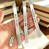 Three stainless steel bookmarks with engraved text and leaf shapes are displayed on a hand against a background of books.