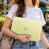 A woman holds a light green matte hard shell case for a MacBook in an outdoor urban setting.