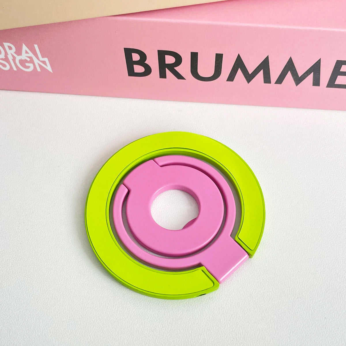 A pink and lime green magnetic phone grip and stand with a foldable ring resting on a white surface.