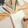 A collection of Morandi Gel Pens in various colors and shades laid out on a wooden desk next to a notebook.